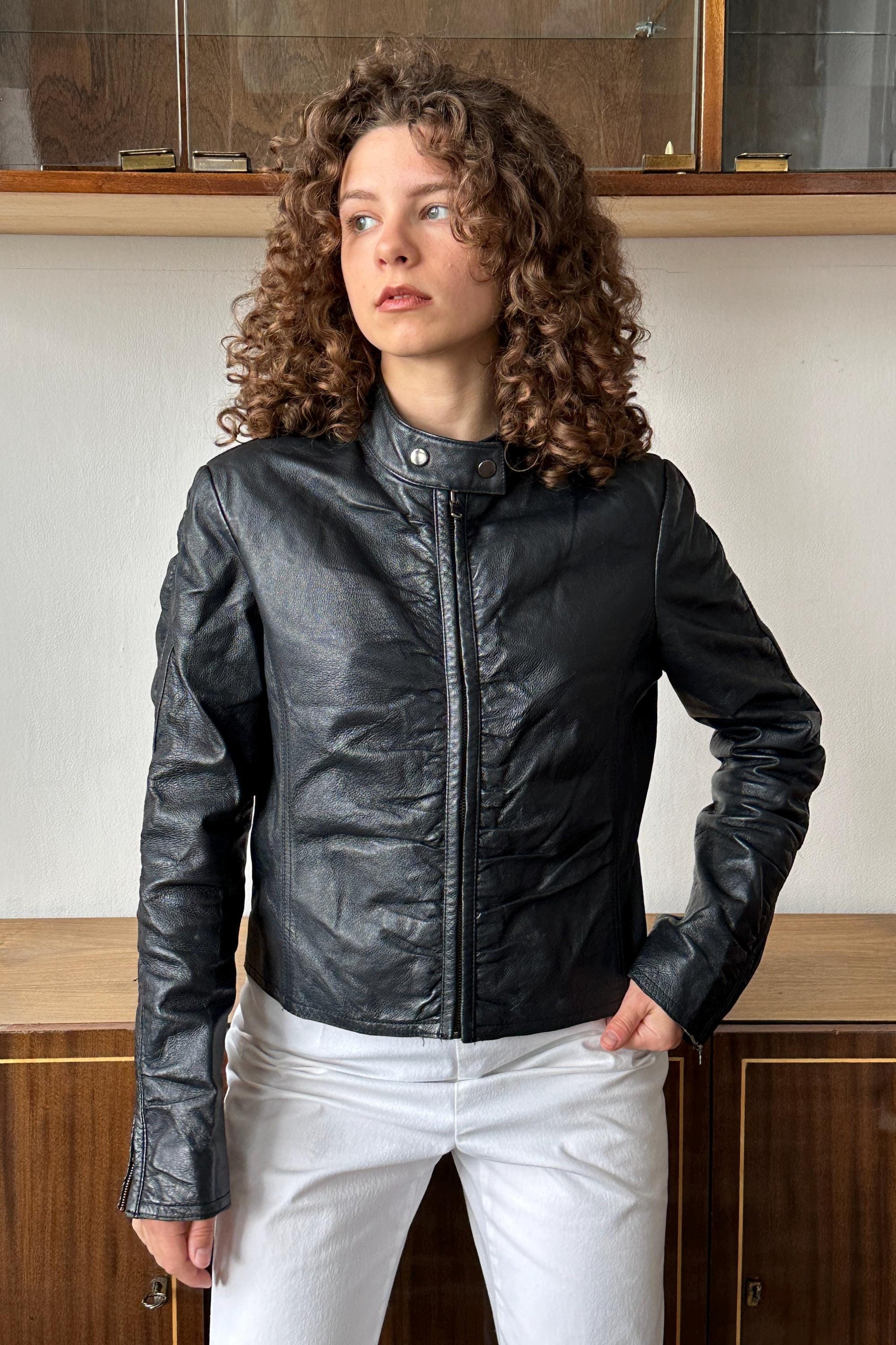 A woman with curly hair wearing a black leather jacket stands in front of a wooden cabinet.