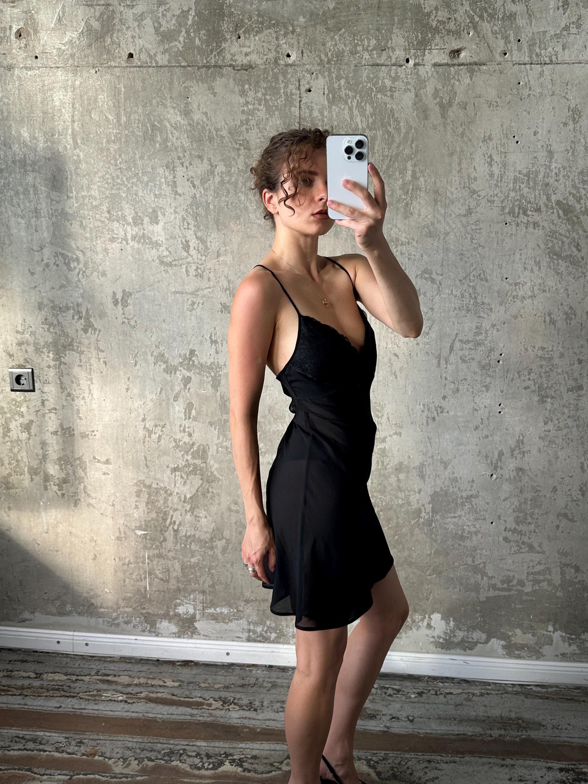 A woman in a black dress is taking a selfie with a white iPhone in front of a concrete wall.