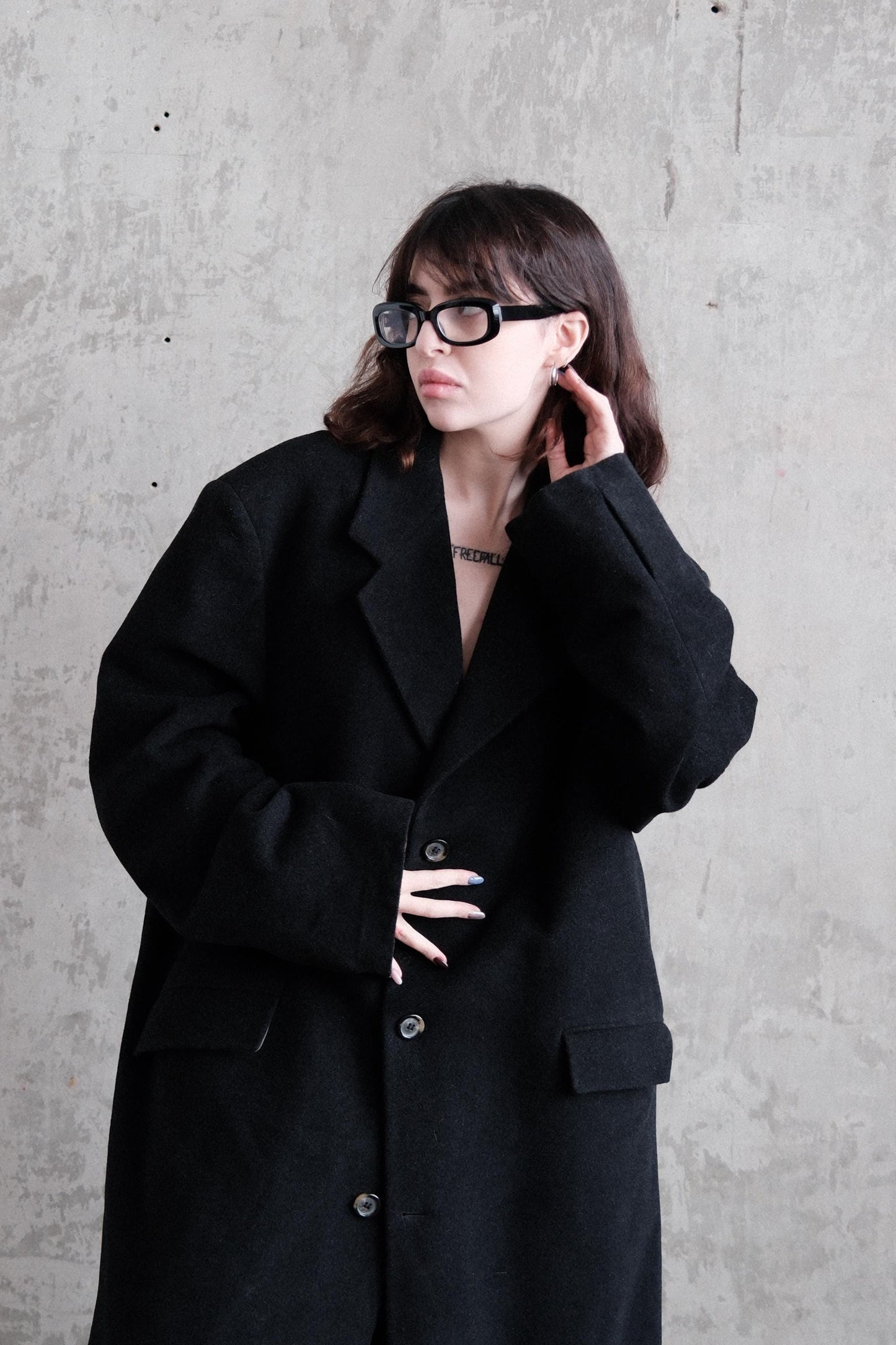 A woman wearing a black coat and glasses is standing against a concrete wall.