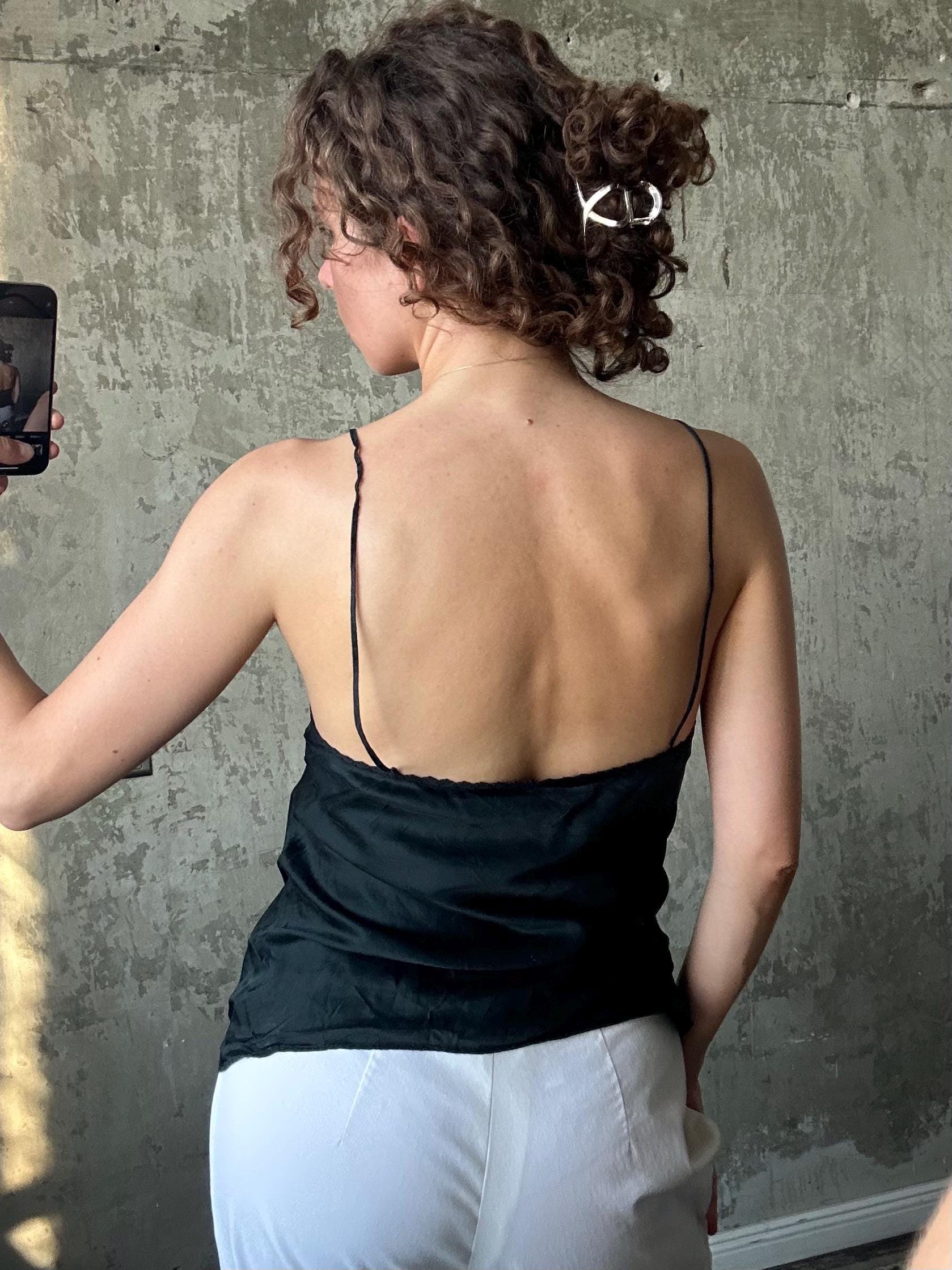 a woman taking a selfie in front of a concrete wall. She is wearing a black top and white pants, and her hair is styled in a bun.