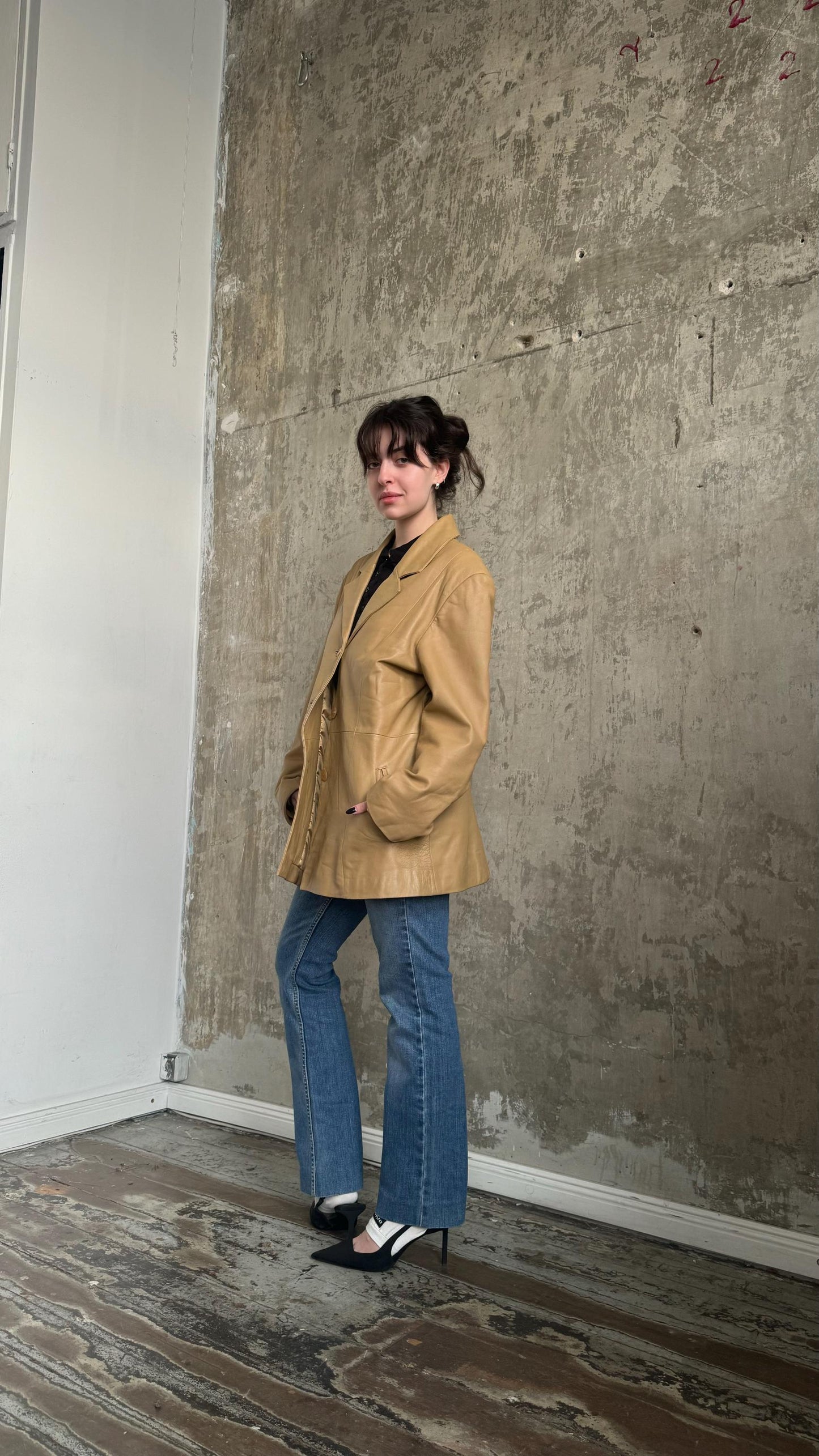 A woman wearing a tan jacket and blue jeans stands in front of a concrete wall, posing for the camera.