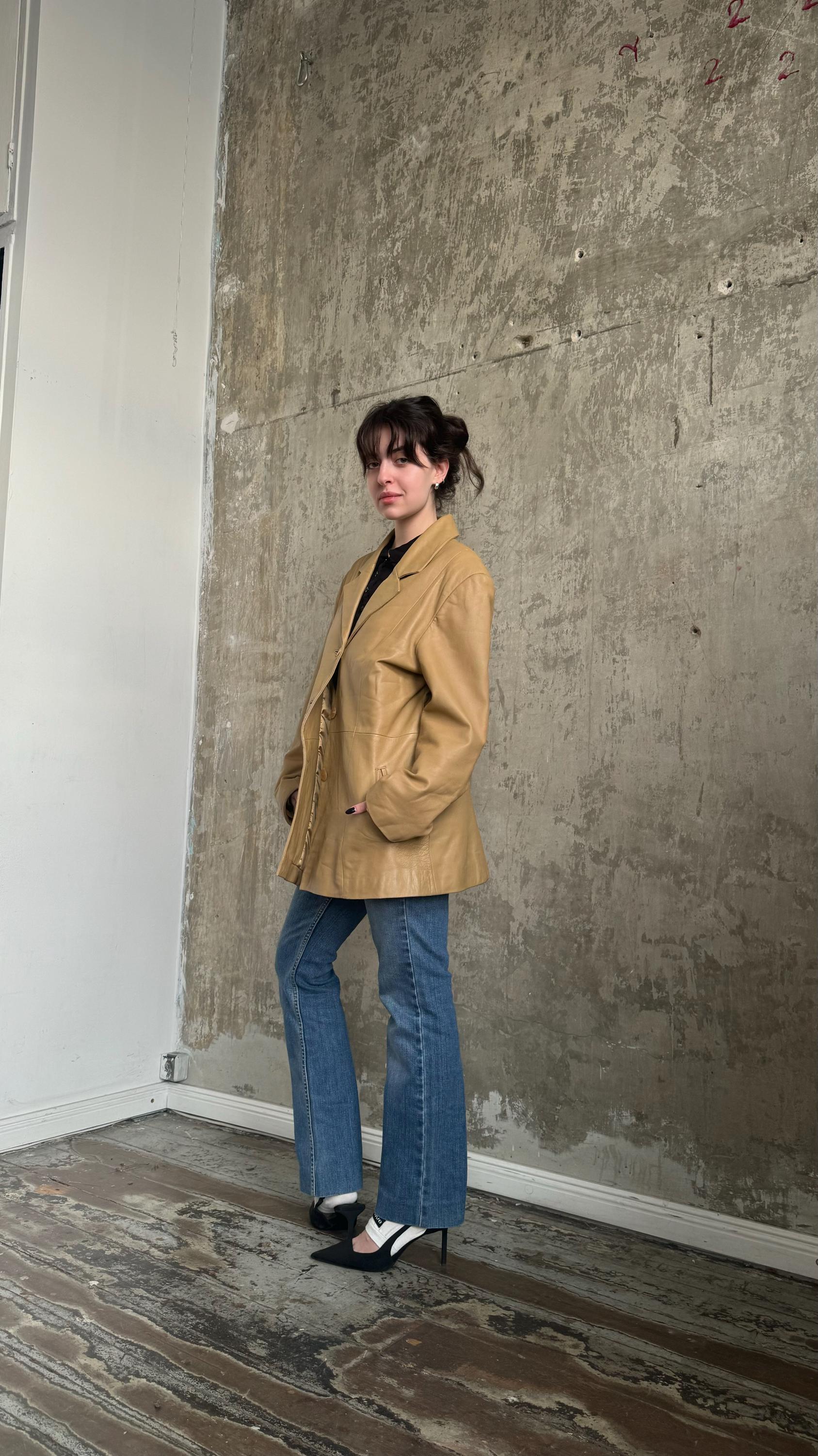 A woman wearing a tan jacket and blue jeans stands in front of a concrete wall, posing for the camera.