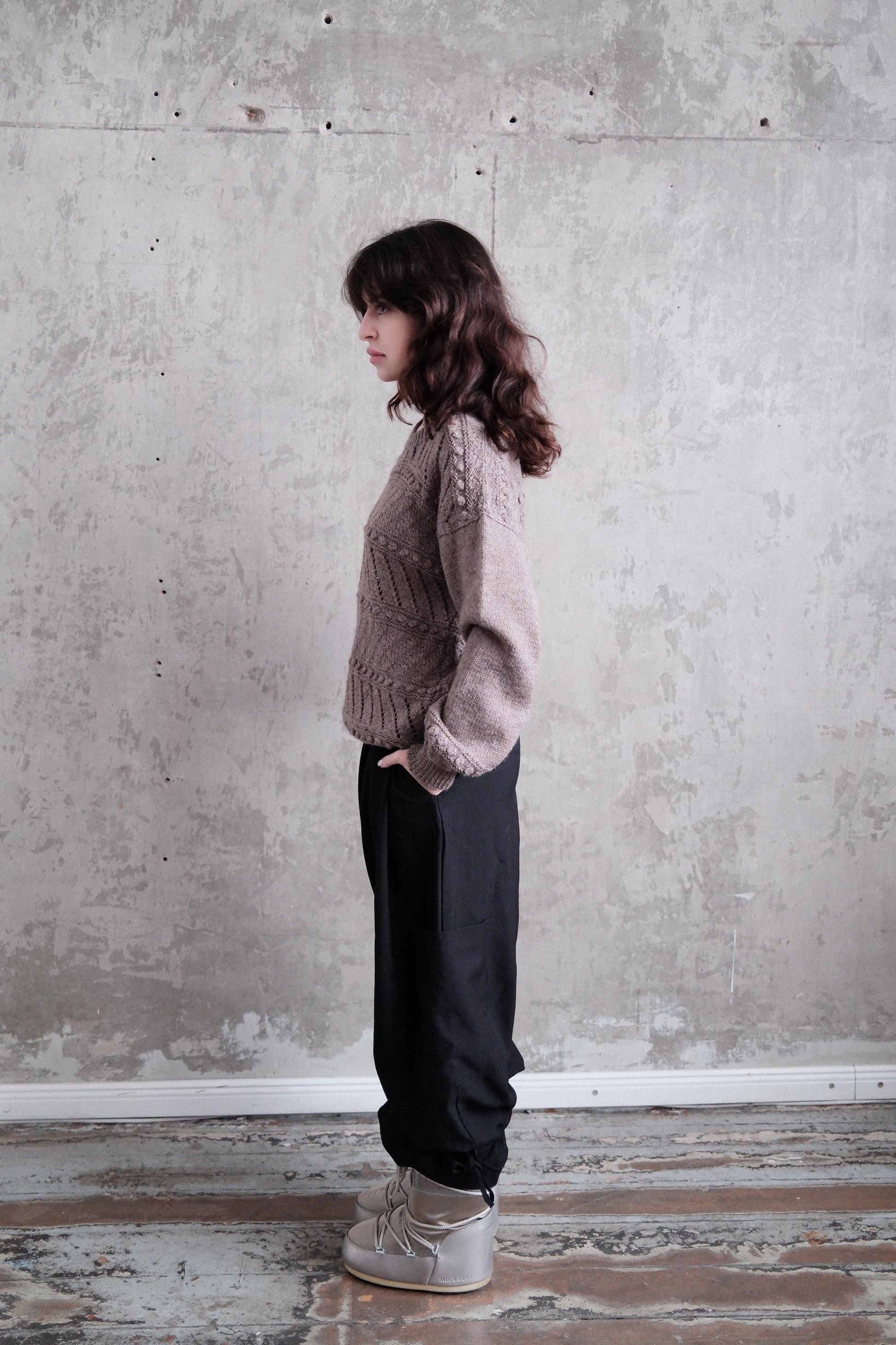 A woman with long brown hair is standing in a room with a concrete wall, wearing a gray sweater and black pants.