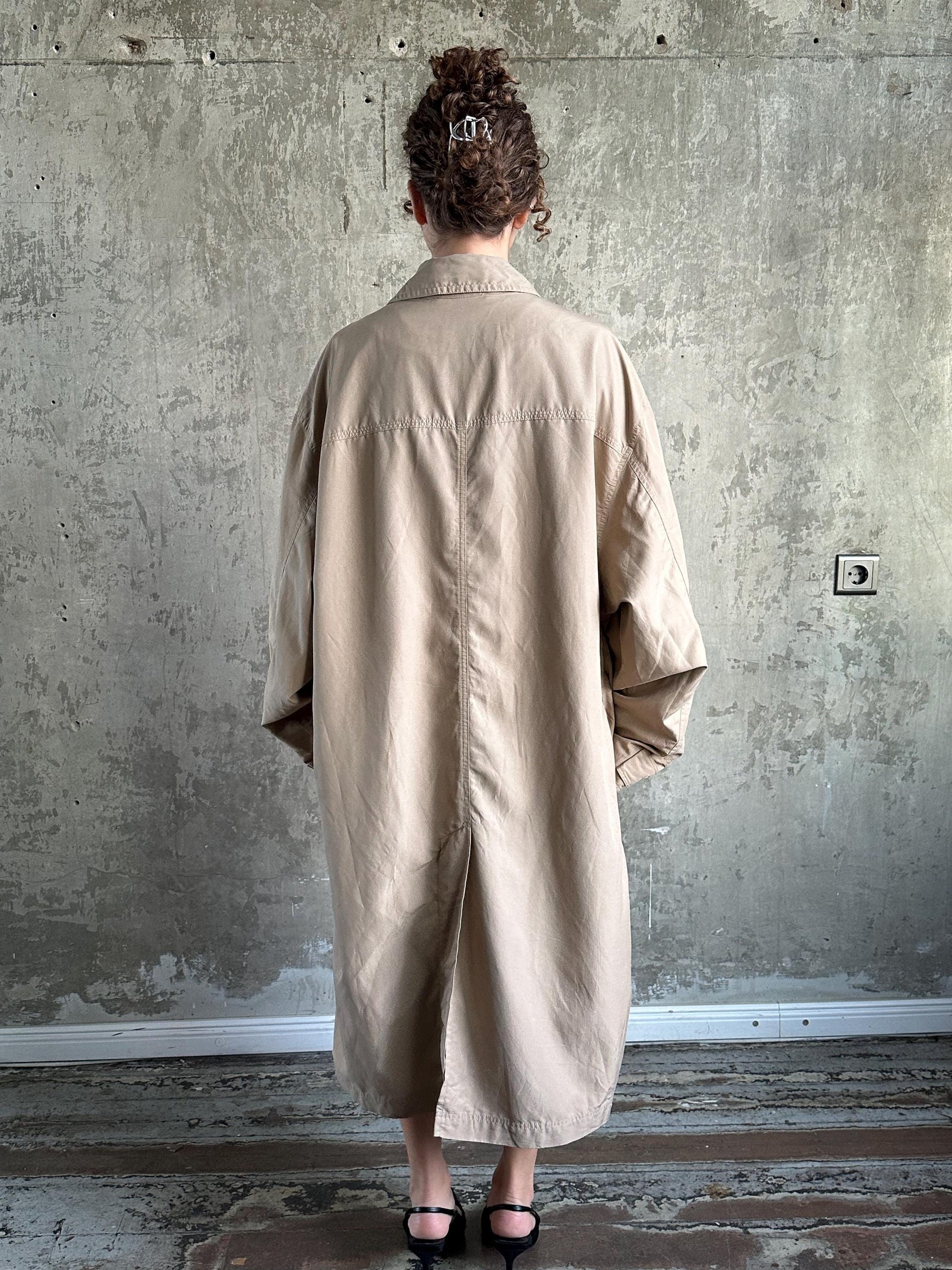 A person wearing a beige coat stands in front of a concrete wall, facing away from the camera.