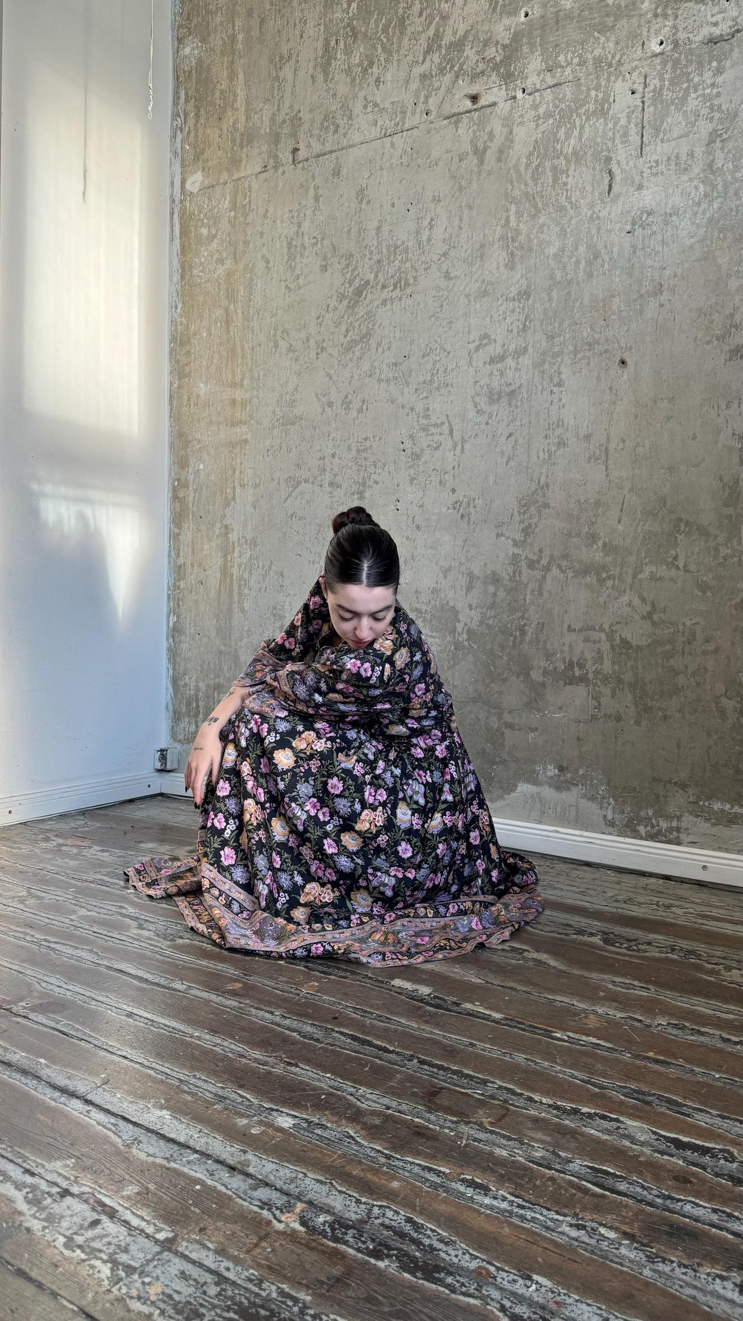A woman in a floral dress sits on a wooden floor, her head resting on her hand, in a room with a concrete wall.