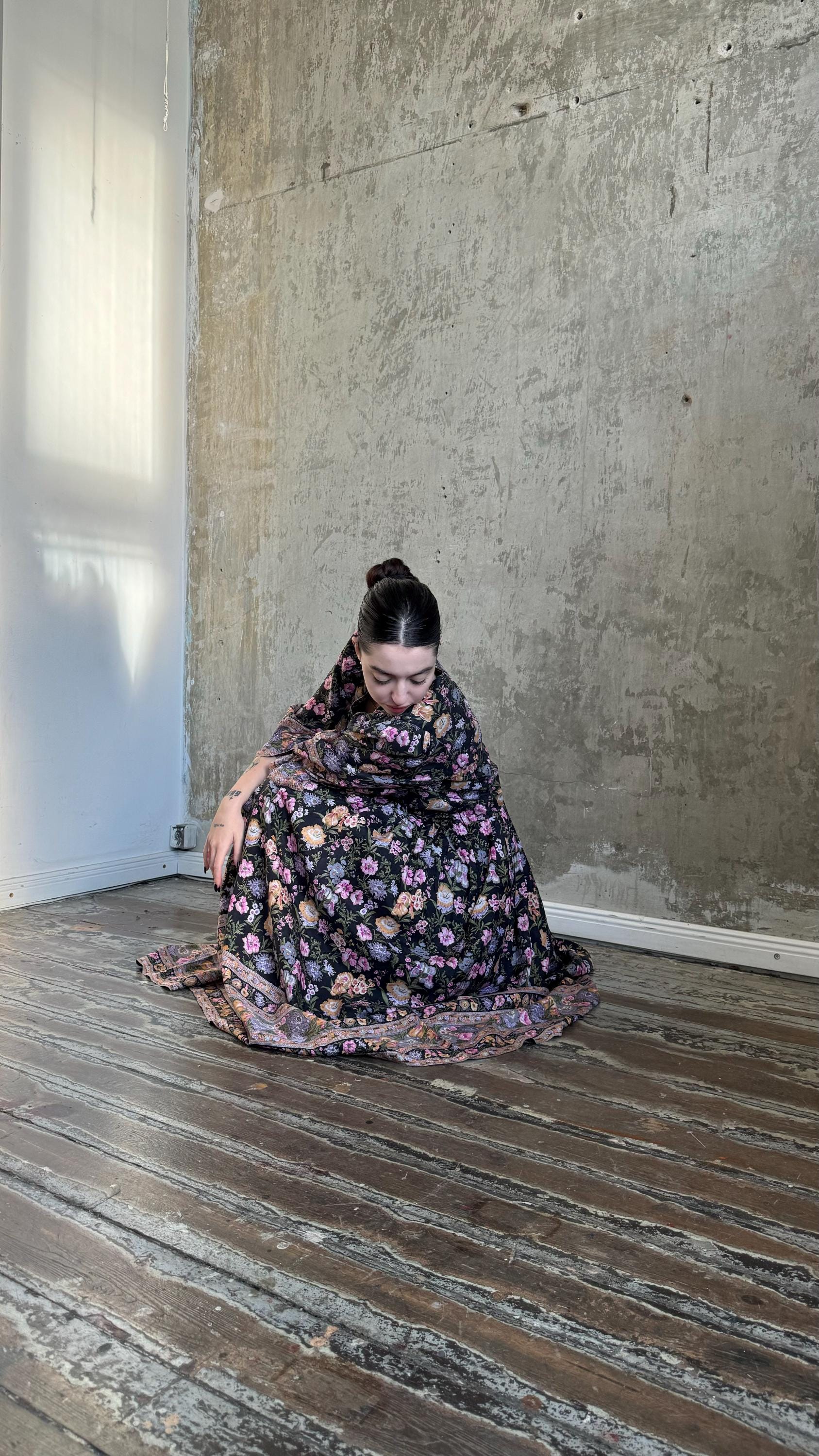 A woman in a floral dress sits on a wooden floor, her head resting on her hand, in a room with a concrete wall.