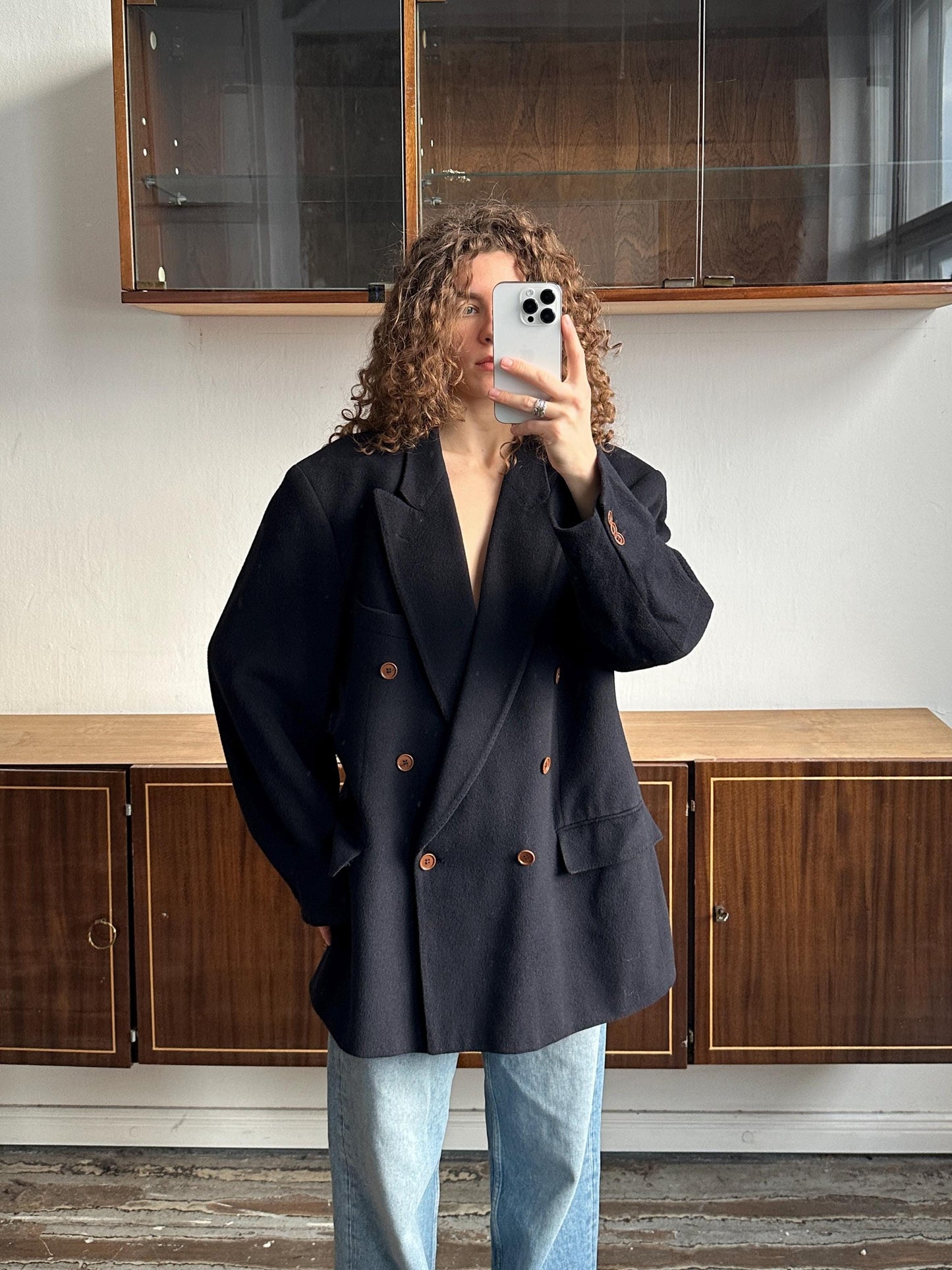 A person with curly hair is taking a selfie with a black blazer and jeans in front of a wooden cabinet.