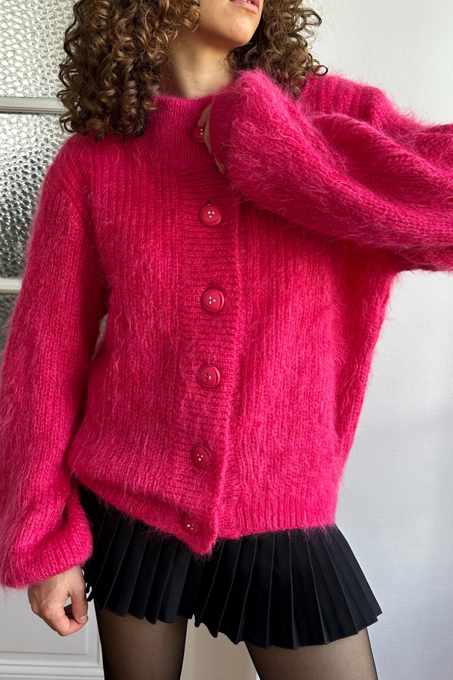 A woman wearing a bright pink cardigan with red buttons and a black pleated skirt.