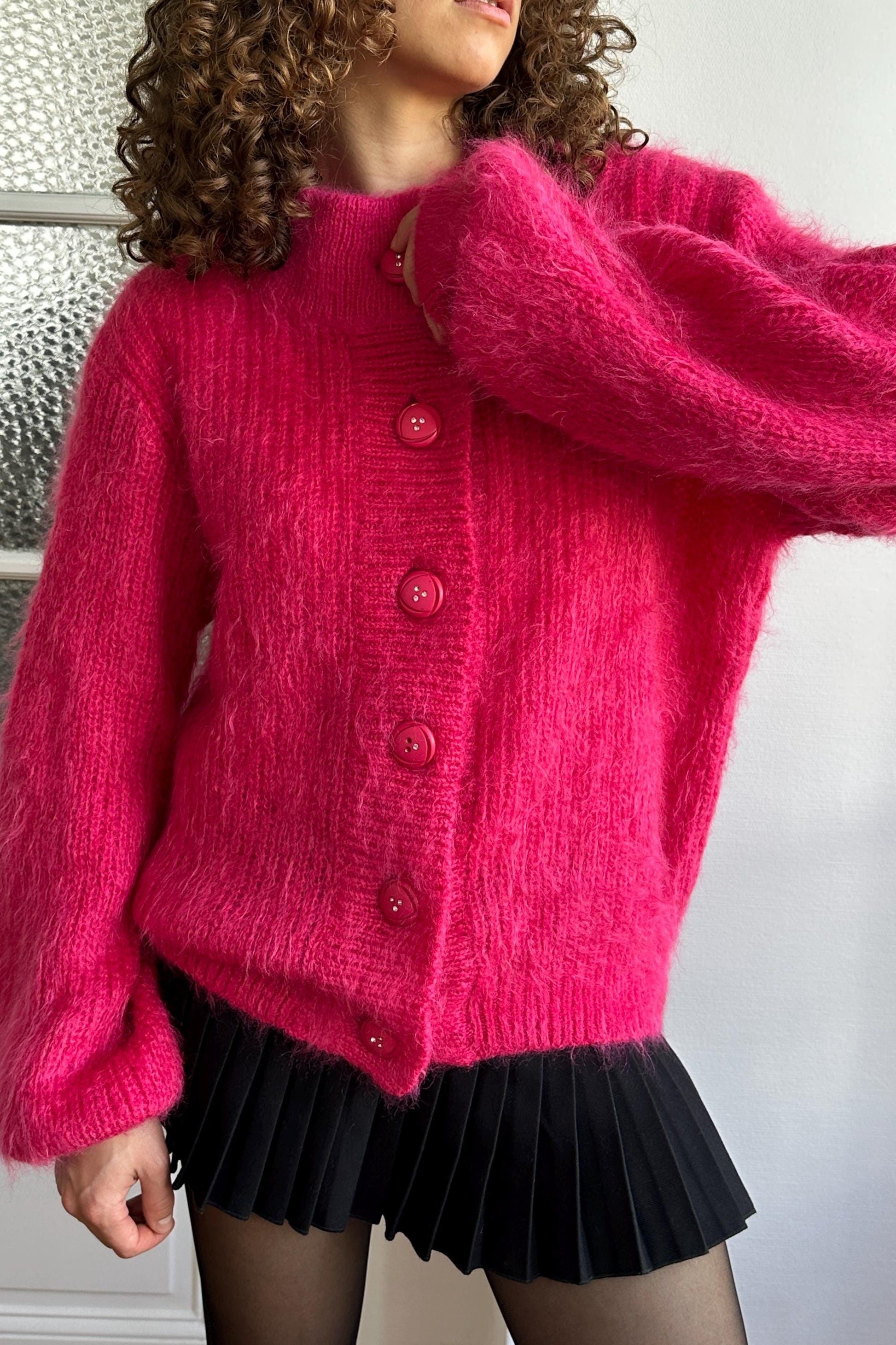 A woman wearing a bright pink cardigan with red buttons and a black pleated skirt.