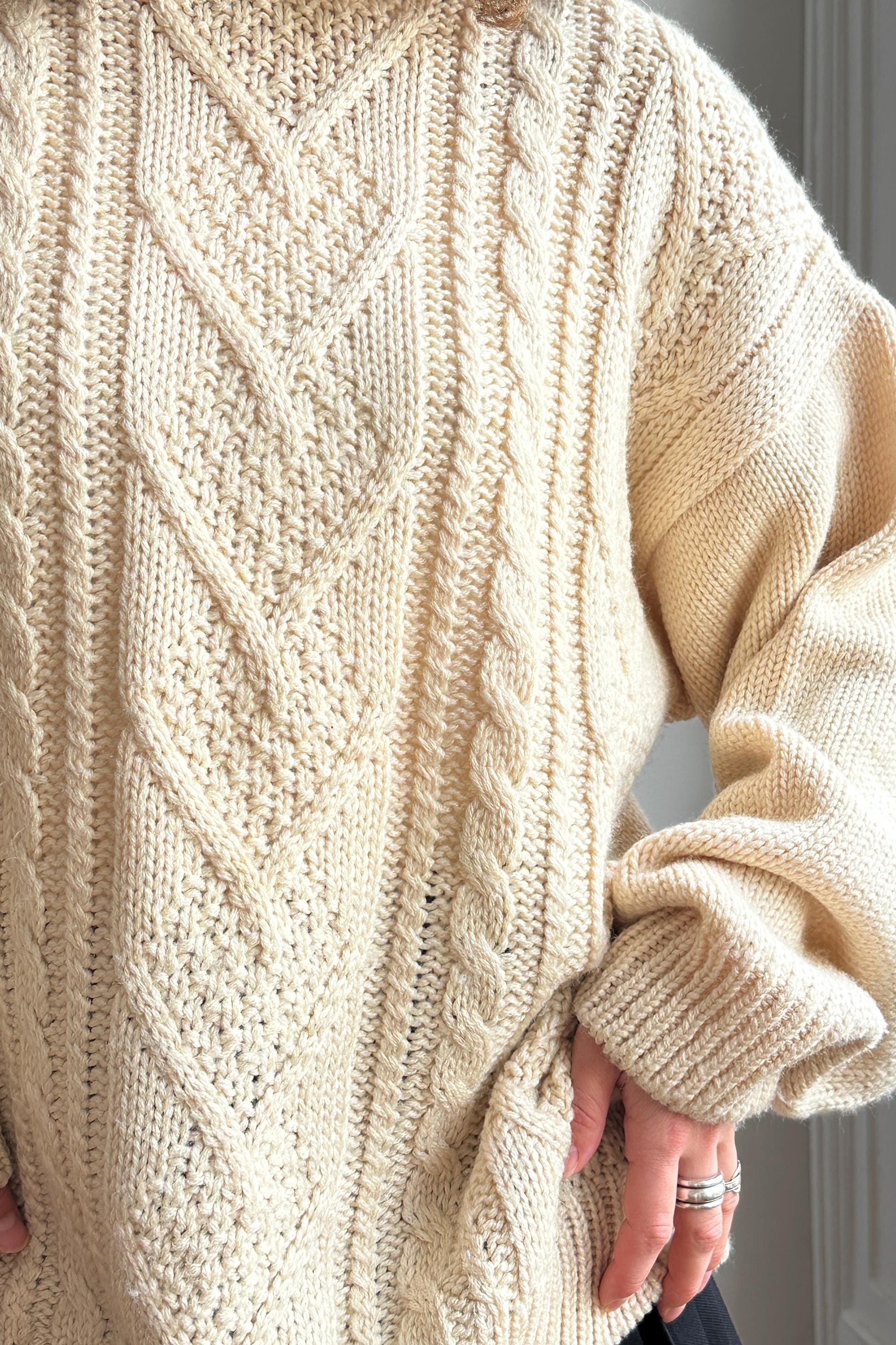 A close-up image of a person wearing a cream-colored knitted sweater with intricate cable patterns.