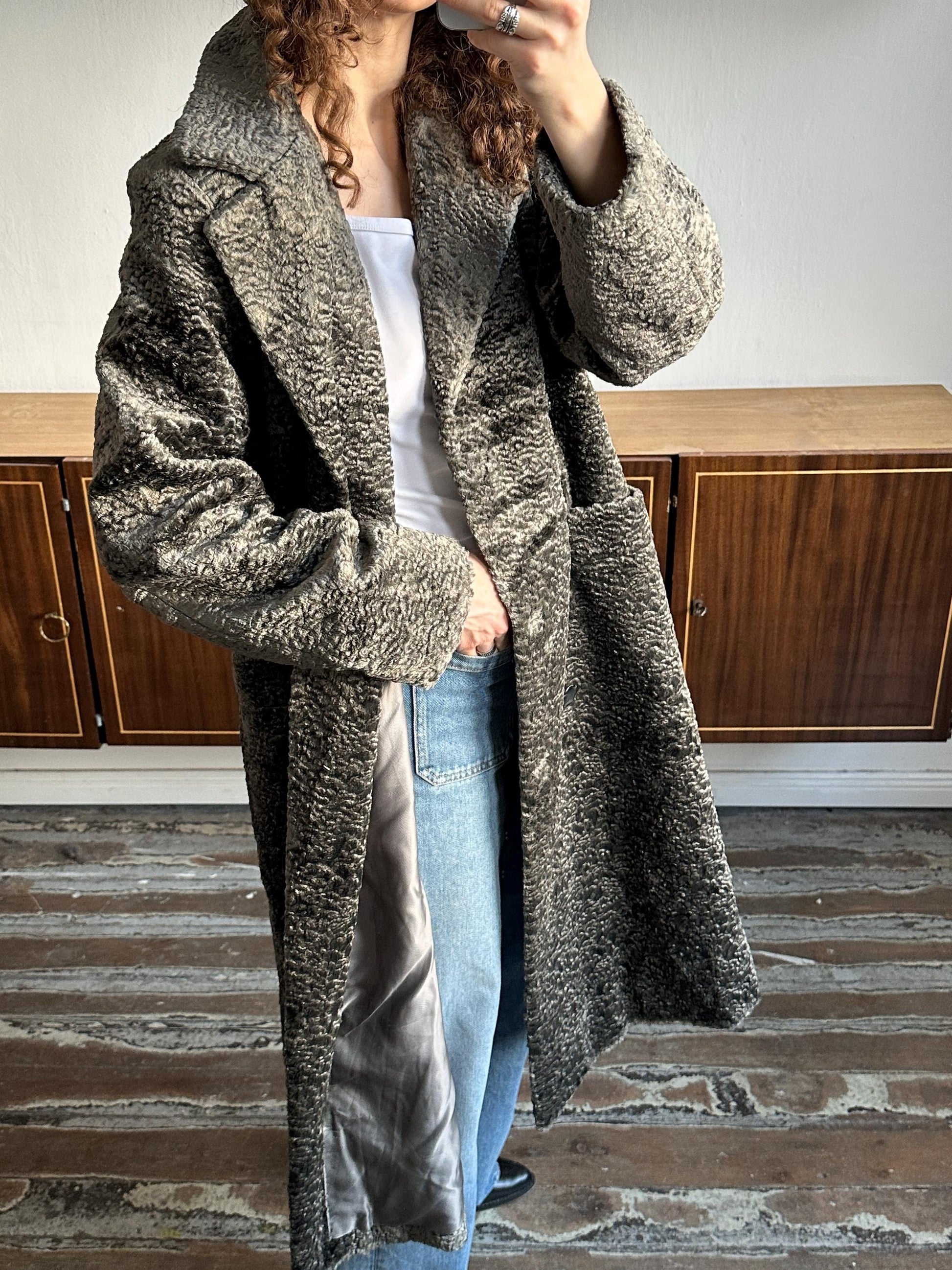 A person wearing a long gray coat and blue jeans is taking a selfie in a room with wooden floors and cabinets in the background.