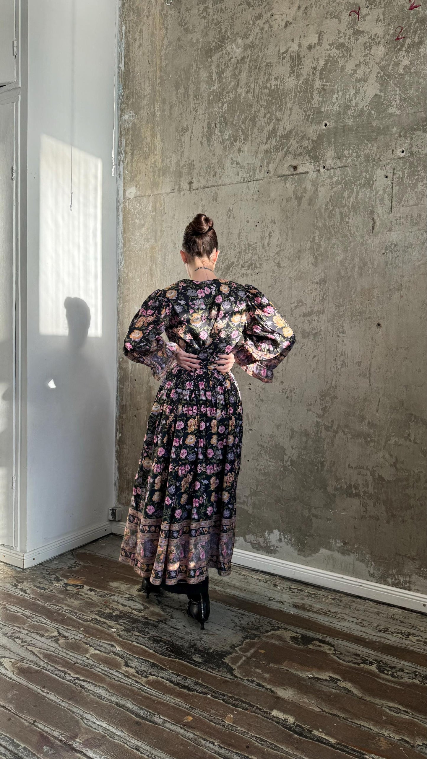 A woman wearing a long, floral-patterned dress stands in a room with a concrete wall and wooden floor.