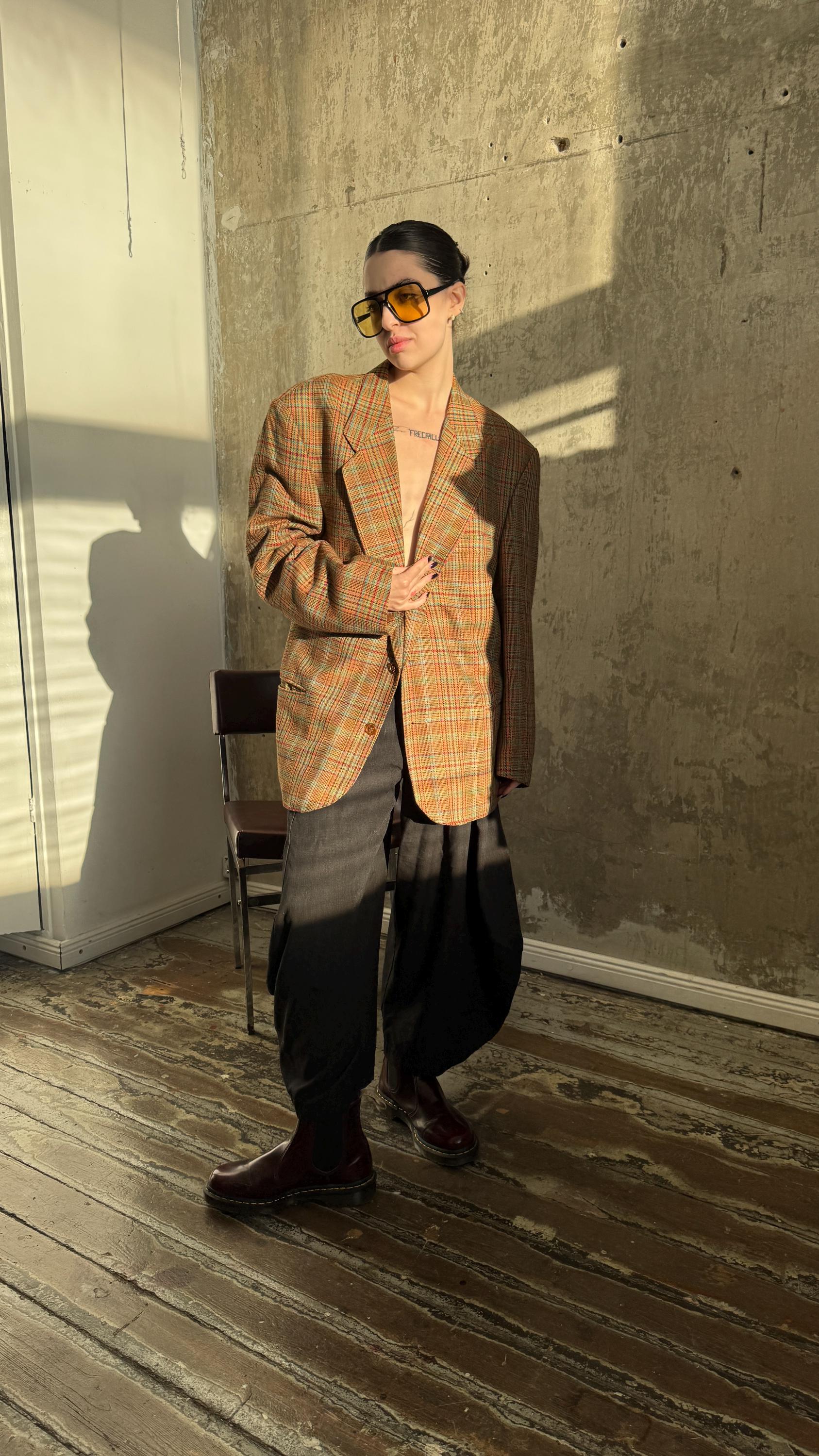 A person wearing a plaid jacket, sunglasses, and standing in a room with a wooden floor and a chair.