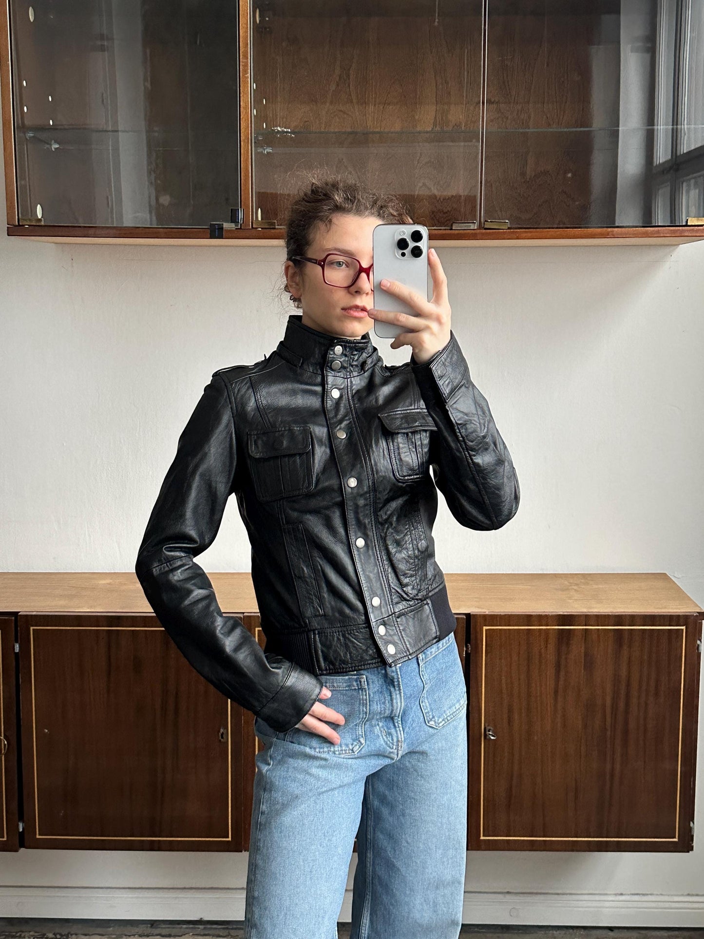 A person wearing a black leather jacket and jeans is taking a selfie with a smartphone in front of a wooden cabinet.