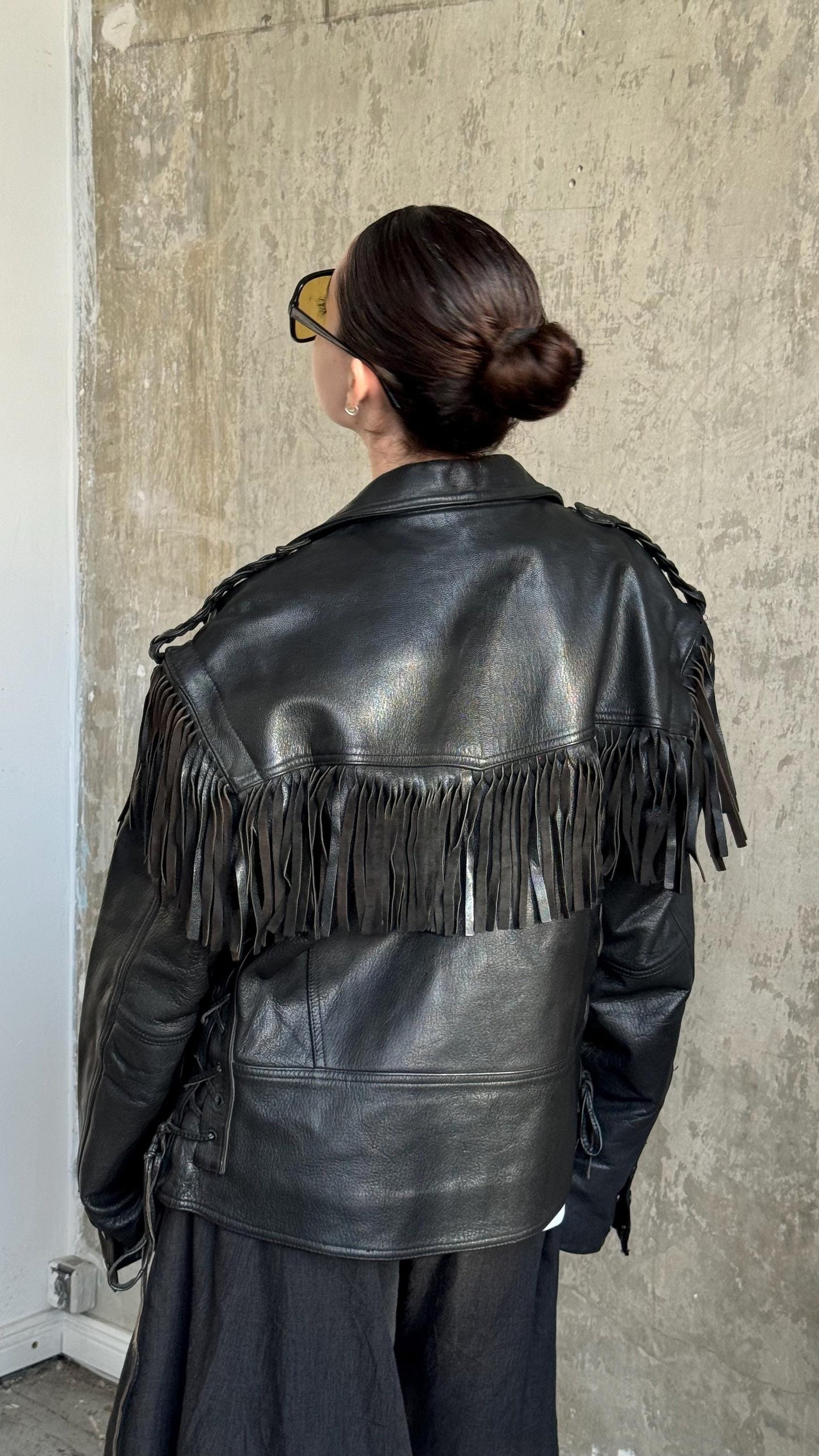 A person wearing a black leather jacket with fringe details, standing in front of a concrete wall.