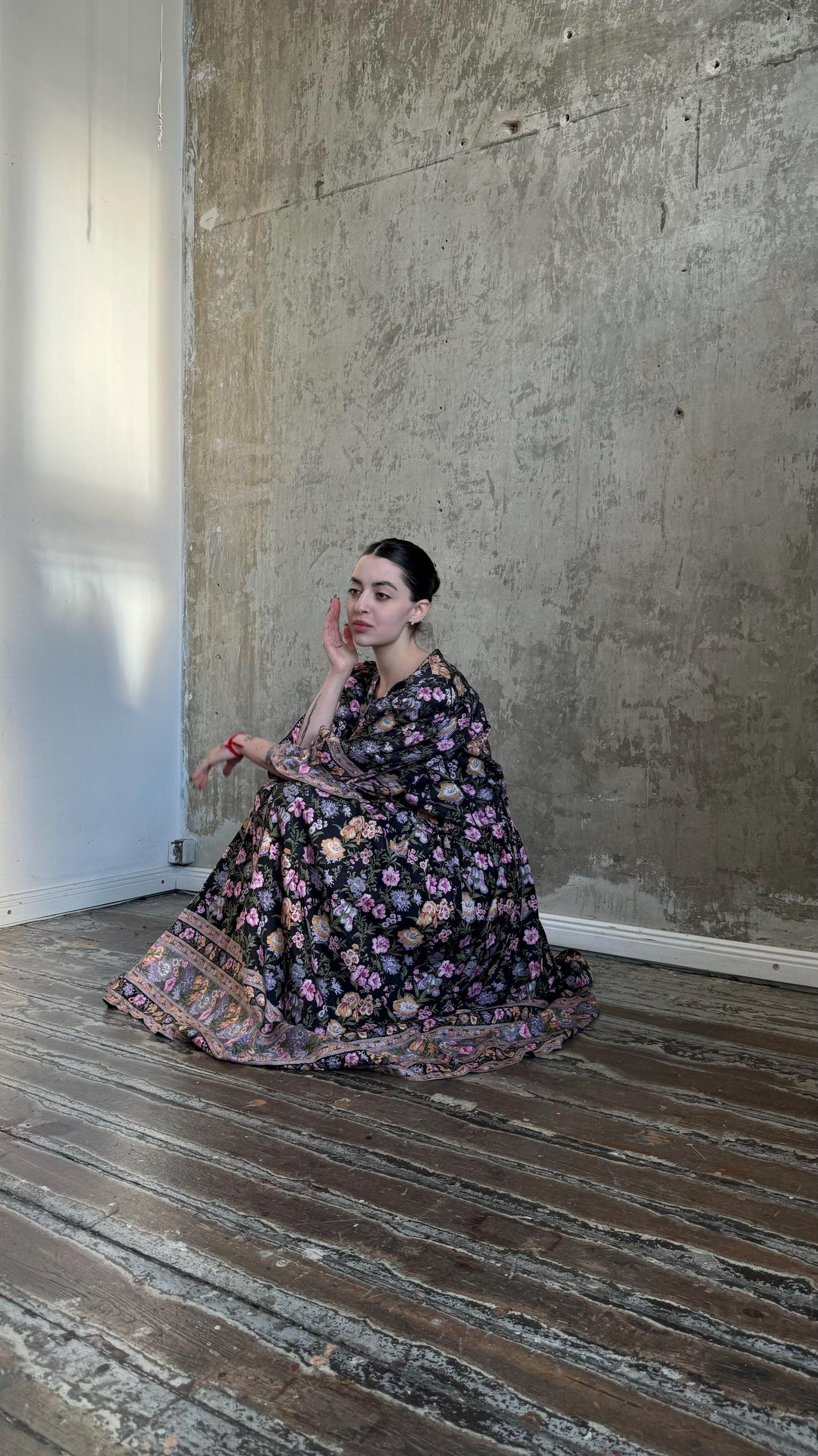 A woman in a long, floral-patterned dress sits on a wooden floor, her hand resting on her chin.